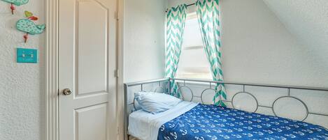 Een eenpersoonsbed met een bedsprei met een patroon, een witte deur en een raam met ruitvormige gordijnen.