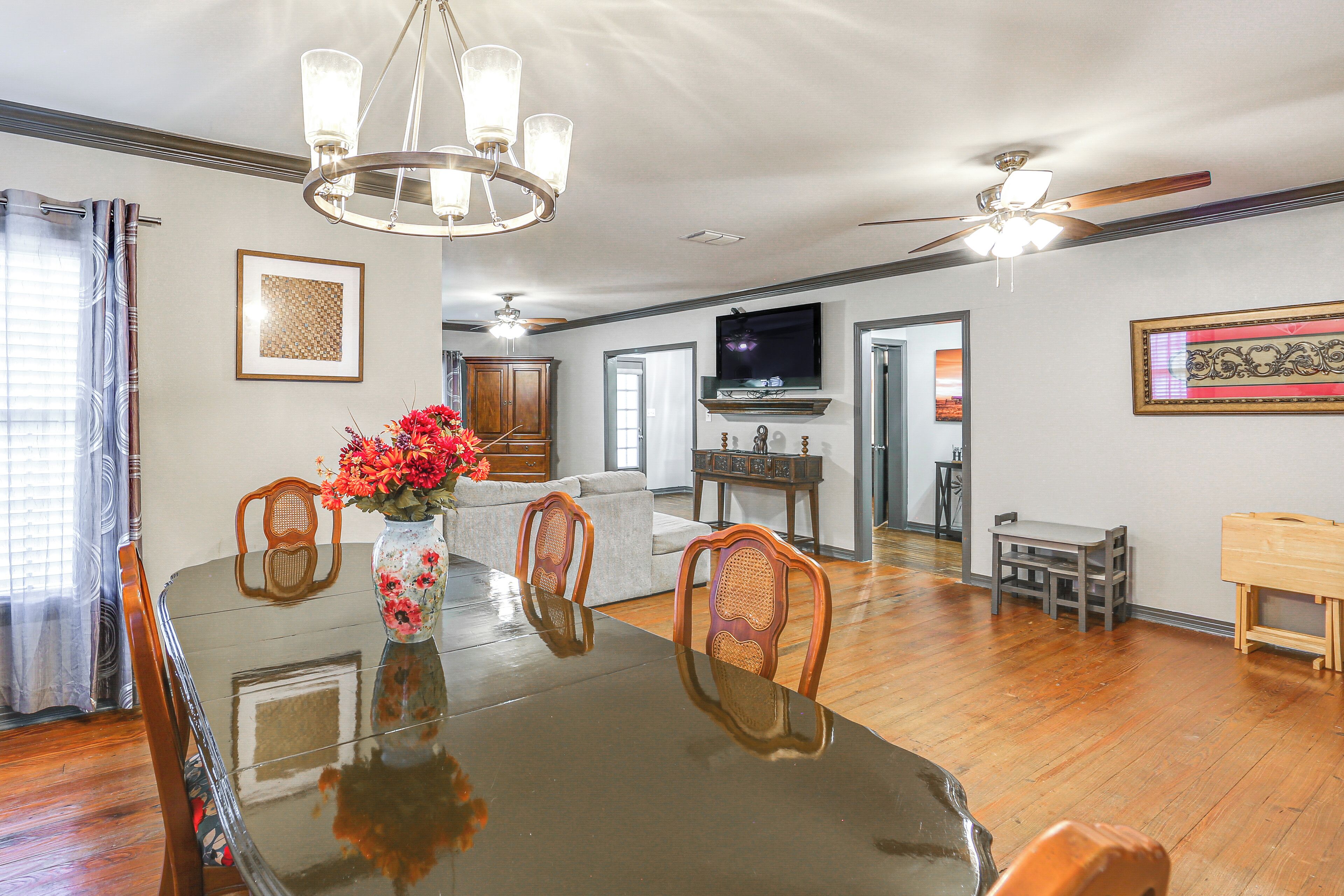 A dining area with a glass table, wooden chairs, a vase with flowers, a ceiling fan, and a TV in the background.