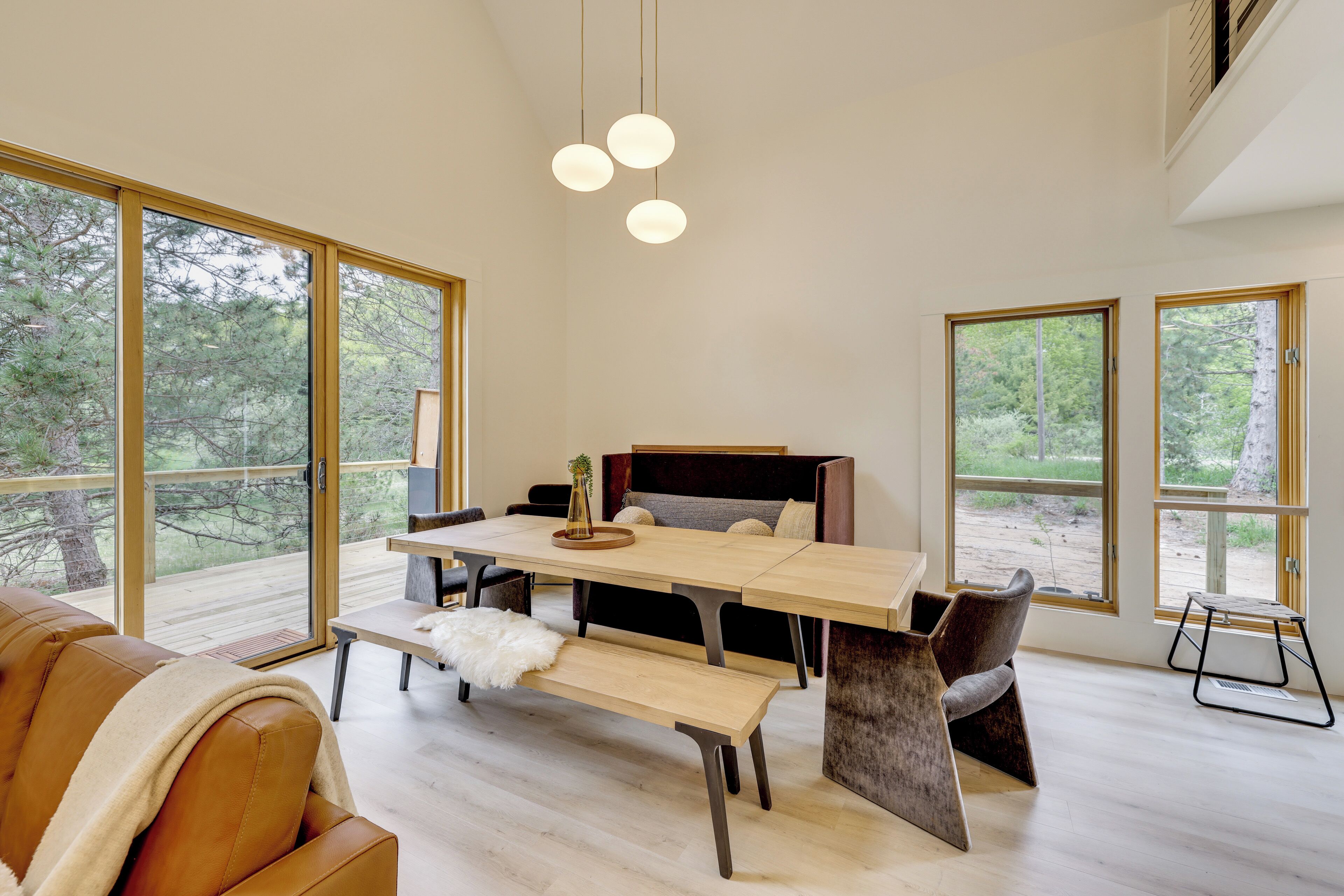 Un interior moderno con una mesa de comedor, un banco y sillas, ventanas amplias con vista a un bosque, y una lámpara de techo.