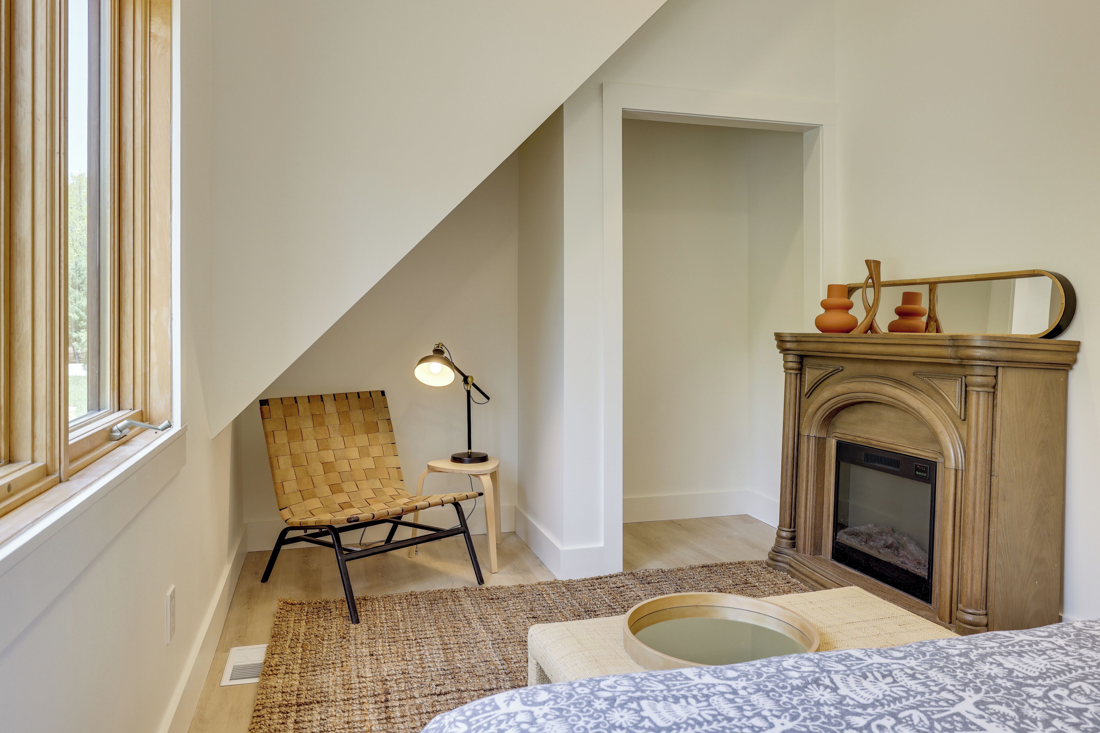 A room with a sloped ceiling, a woven chair, a small table with a lamp, a fireplace, and a bed with patterned bedding.