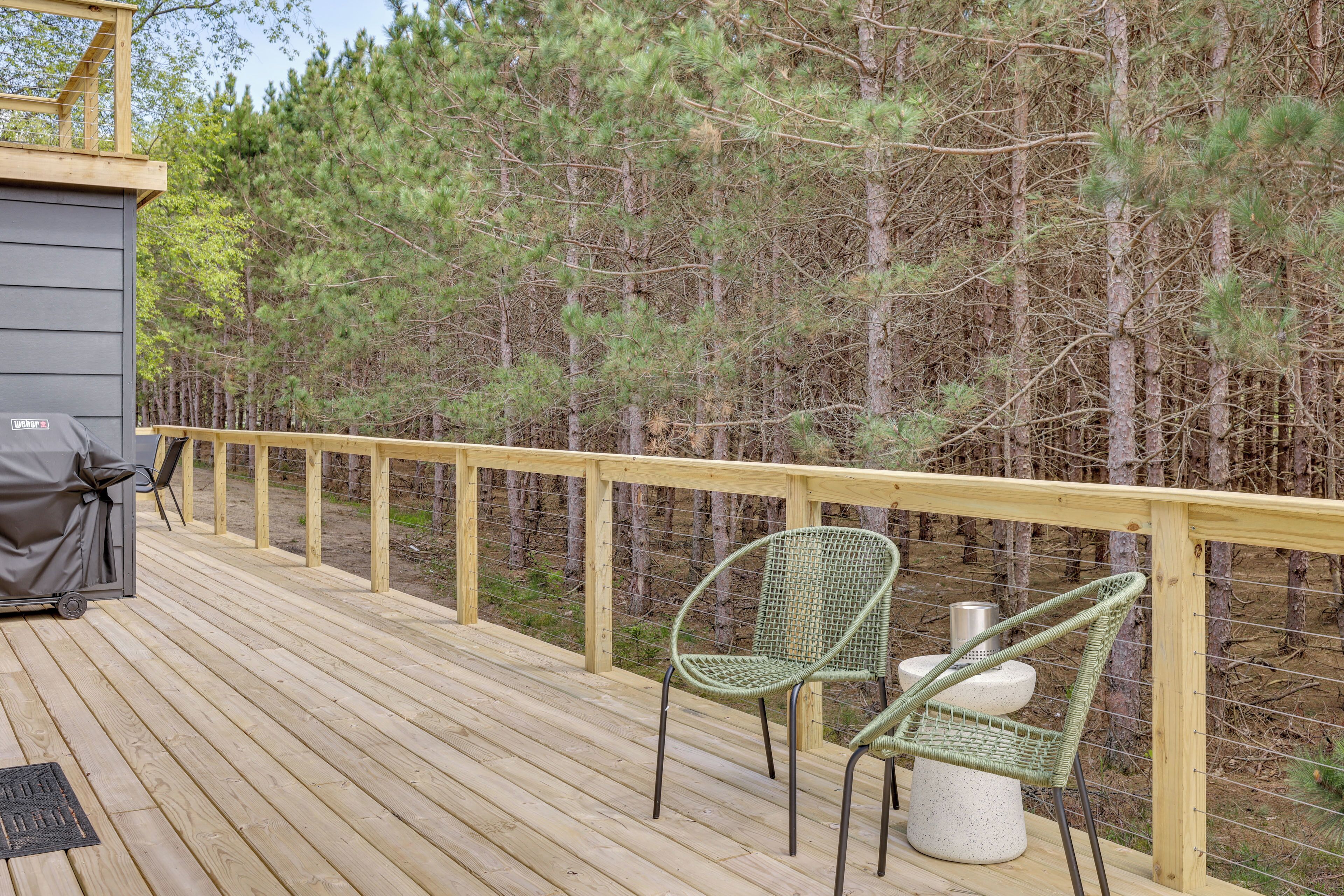A wooden deck with two green chairs and a grill, surrounded by a forest.