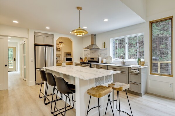 A modern kitchen with a large island, bar stools, stainless steel appliances, and a view of trees through a window.