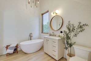 Un baño moderno con una bañera independiente, un tocador con cajones, un espejo redondo y una planta en maceta.