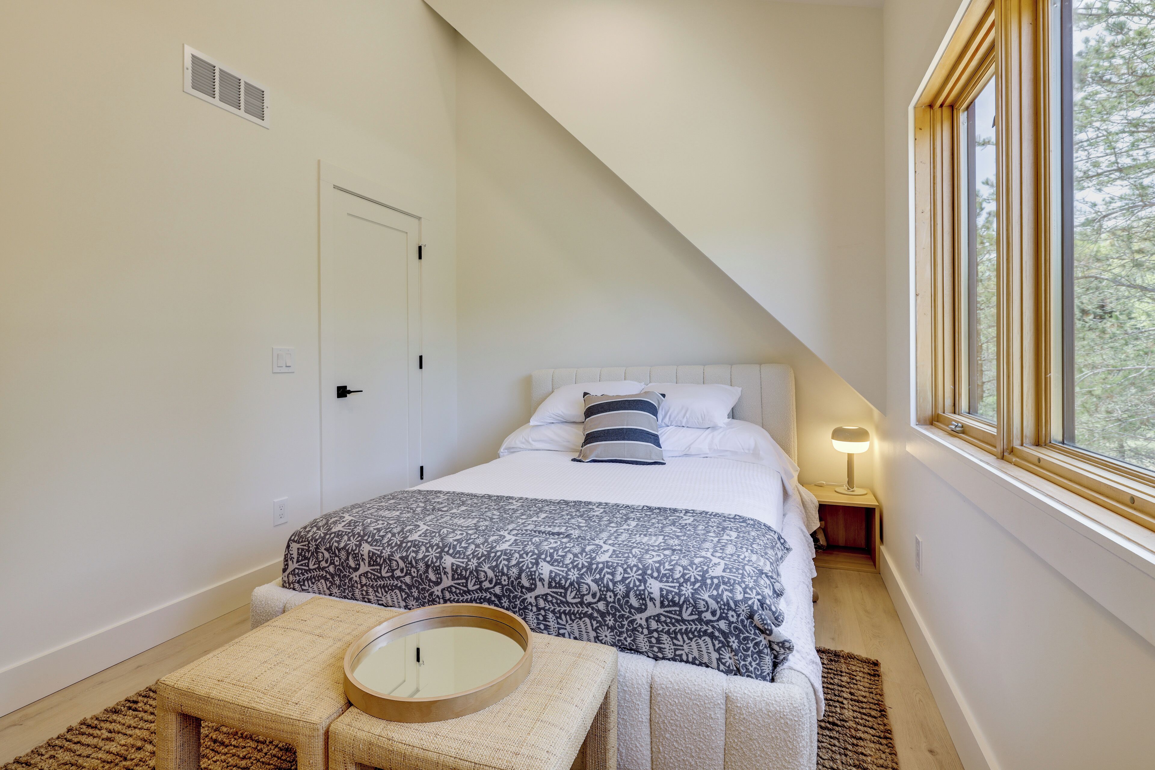 A bedroom with a bed, bedside table, lamp, and a window with a view.