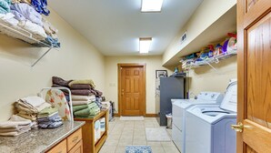 Un lavadero con lavadora, secadora, estantes con productos de limpieza, una zona de planchado y una puerta que da a otra habitación.