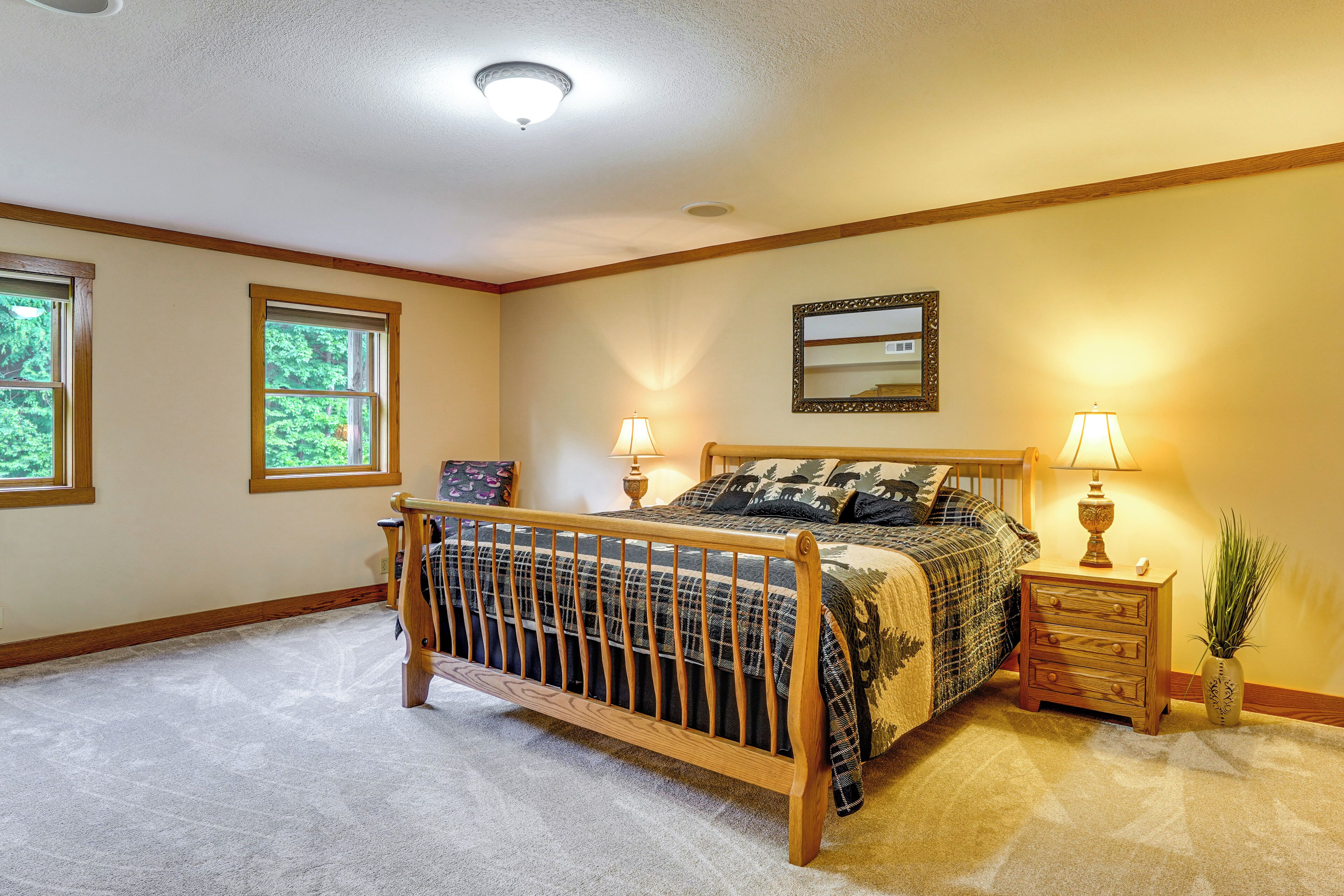 A bedroom with a wooden bed, two bedside lamps, a nightstand, a mirror, and two windows.