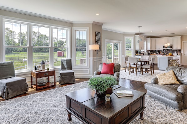 A spacious living room with large windows, a kitchen with white cabinets, and a dining area with a red accent pillow.