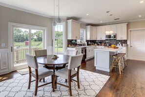A kitchen with white cabinets, a dining area with a round table and chairs, and a view of a green landscape through large windows.