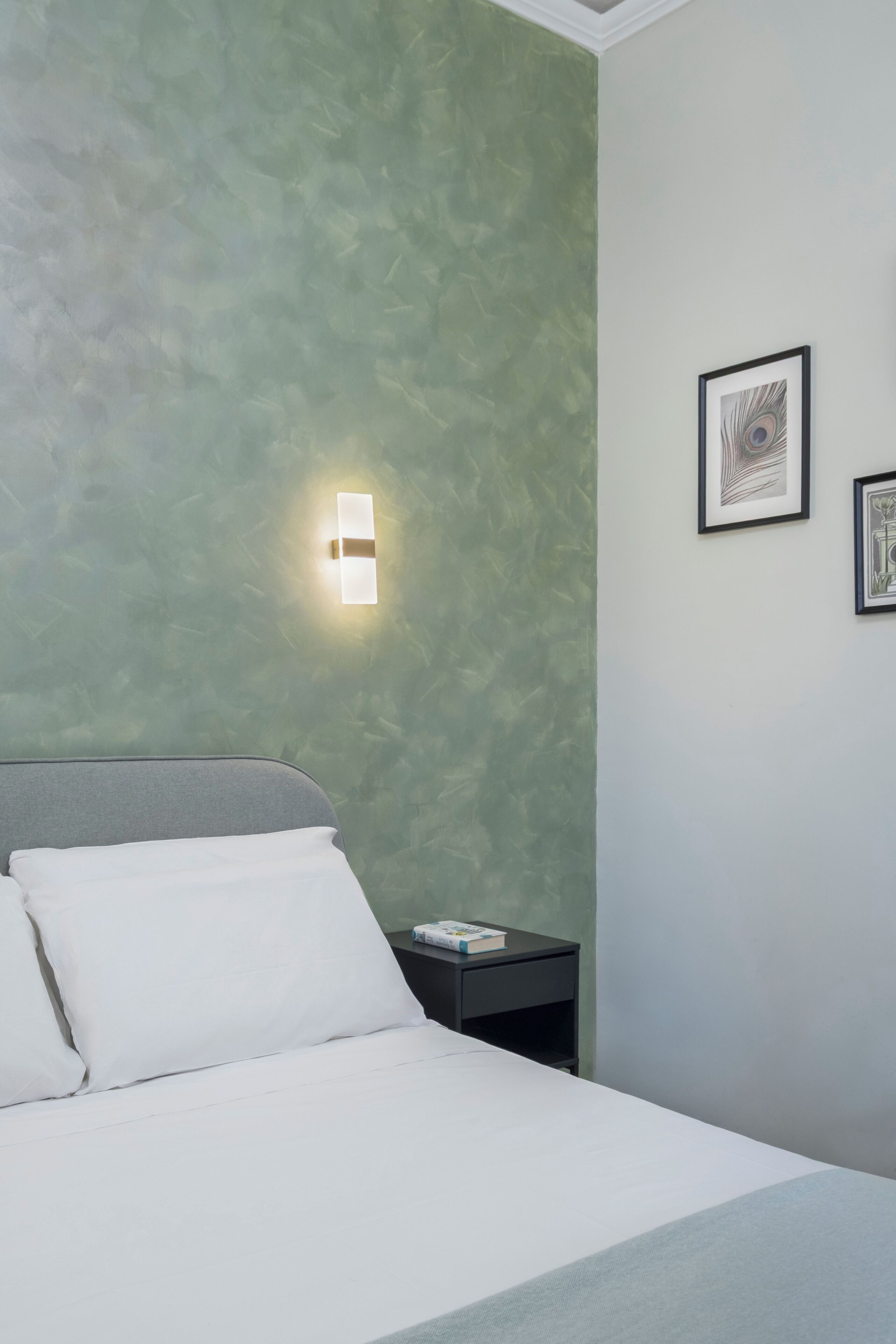 Et hotelværelse med en seng, et natbord, en vægmonteret lampe og to billeder i rammer.
