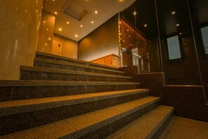 Un escalier en granit mène à une plateforme en bois, avec un mur de verre sur la droite et une porte en haut.