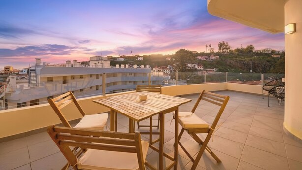 A balcony with a wooden table and chairs, overlooking a cityscape during sunset. - Little Paradise - Funchal (Funchal)