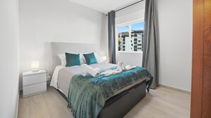 A modern bedroom with a bed, a nightstand, a window with curtains, and a view of buildings outside. - Little Paradise - Funchal (Funchal)
