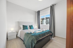 A modern bedroom with a bed, a nightstand, a window with curtains, and a view of buildings outside. - Little Paradise - Funchal (Funchal)