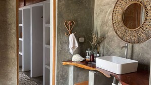 Un baño con pared de concreto, tocador de madera, espejo redondo, lavamanos blanco e iluminación colgante.