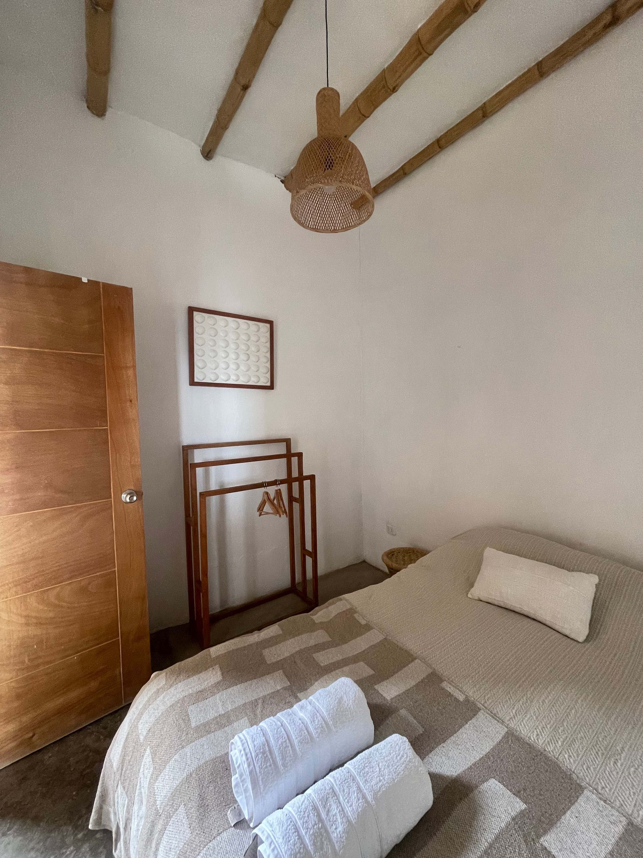 A bedroom with a wooden door, a bed with a patterned bedspread, a hanging light fixture, and a wall-mounted picture frame.