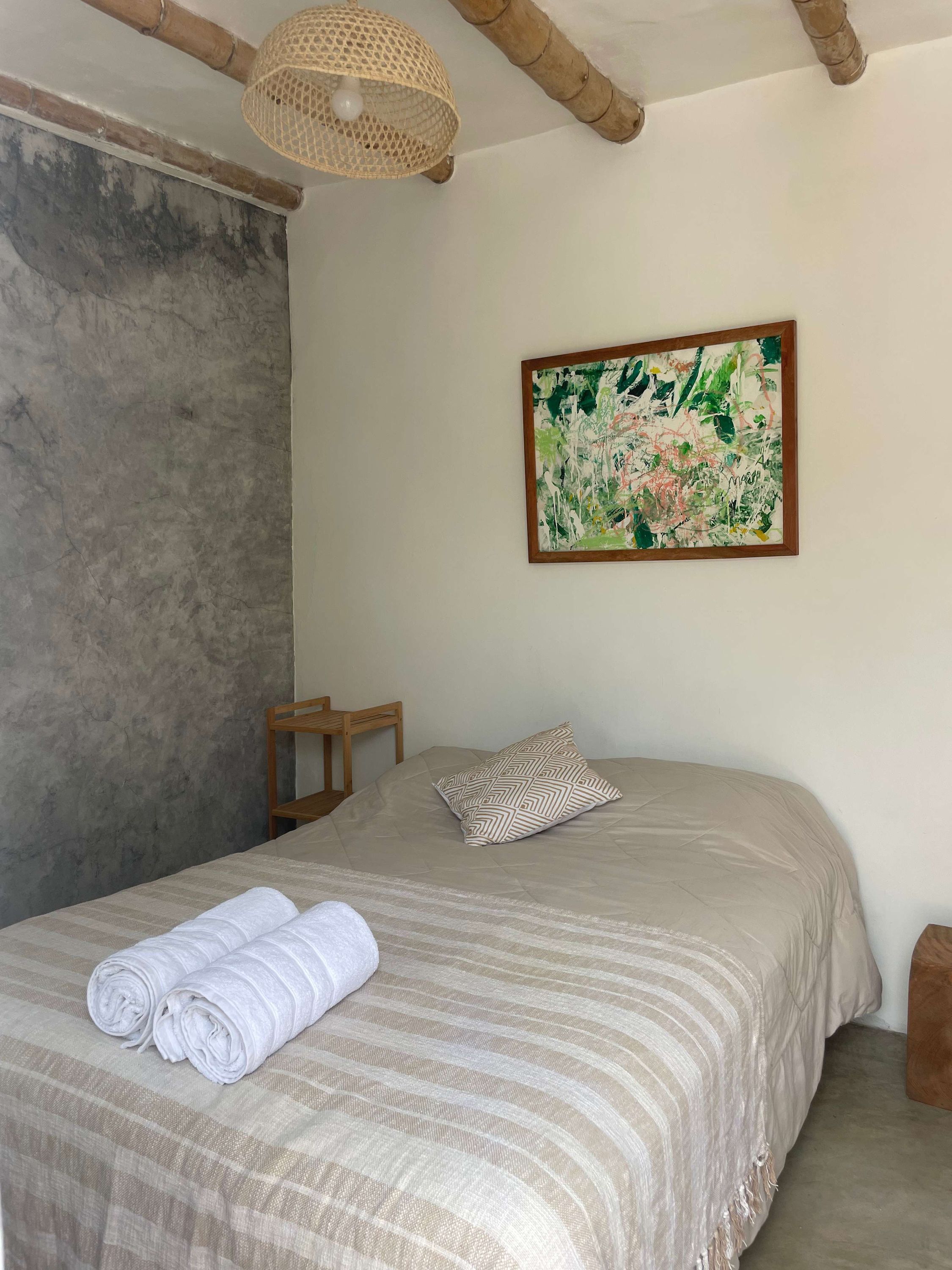 A bedroom with a bed, a hanging light fixture, a wooden chair, a pillow, and a painting on the wall.