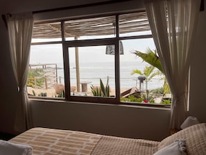 A bedroom with a large window overlooking a beach, a bed, and a view of the ocean.