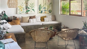Una habitación con techo de paja, un sofá con cojines, sillas de mimbre, una mesa con un florero y una ventana con vistas a la vegetación.