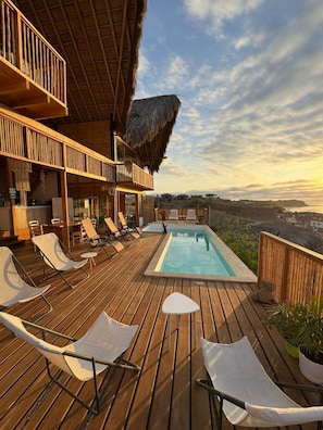 A wooden deck with lounge chairs, a pool, and a thatched roof structure.