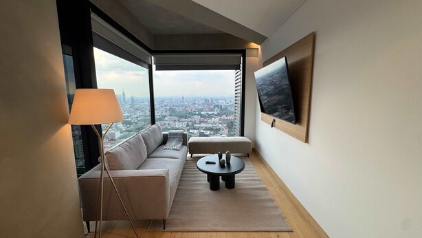 Ein modernes Wohnzimmer mit einer Couch, einem kleinen Tisch, einer Lampe und einem großen Fenster mit Blick auf die Stadt.