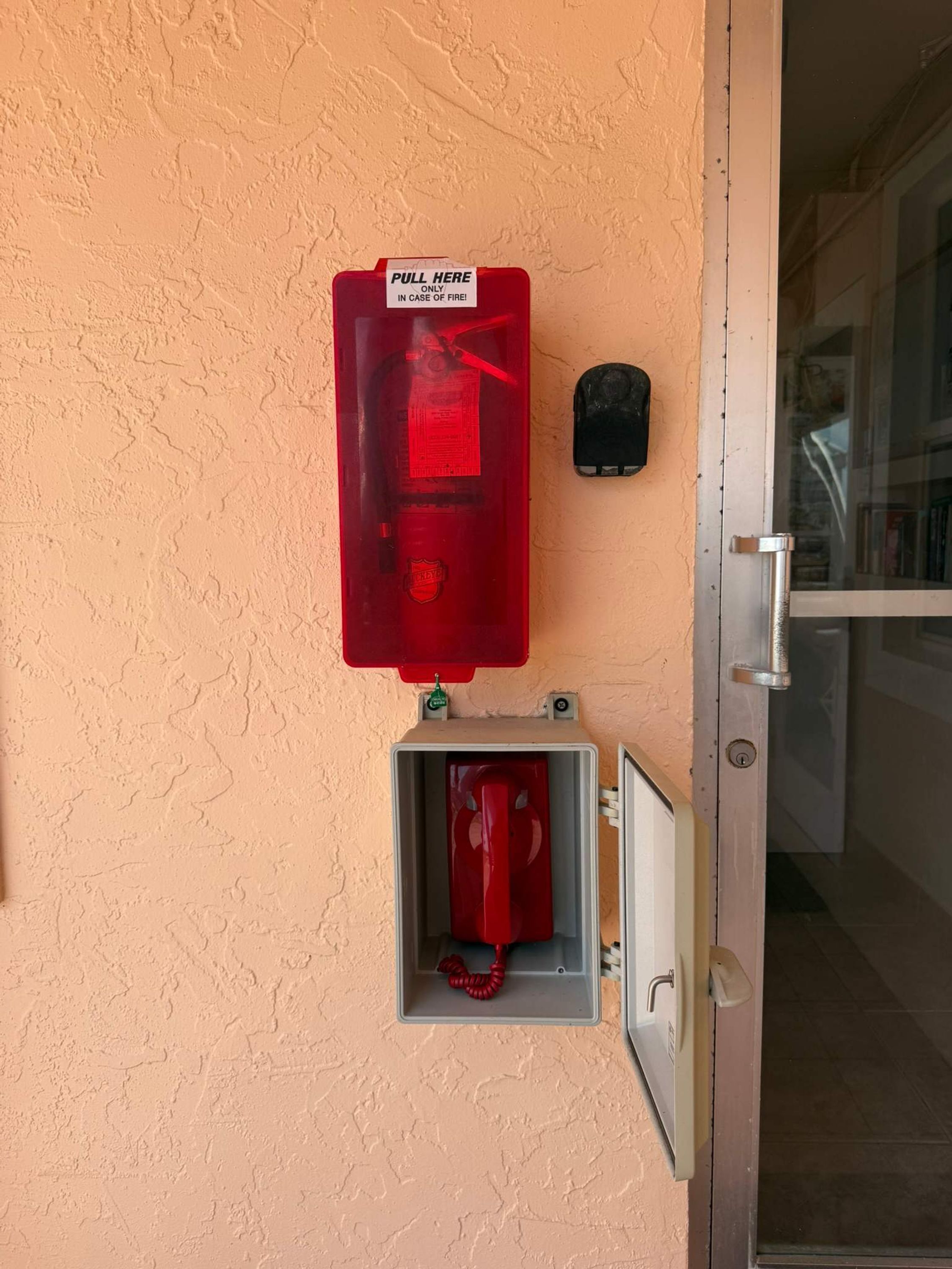 A fire alarm pull station mounted on a textured wall, a door with a handle, and a small, open electrical box with a red cable.
