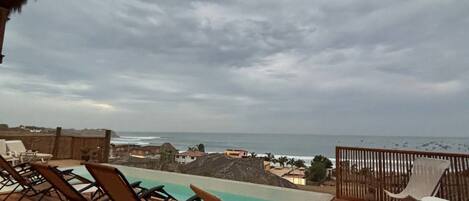 A wooden deck with lounge chairs and a small pool, overlooking a coastal area with buildings and the ocean.