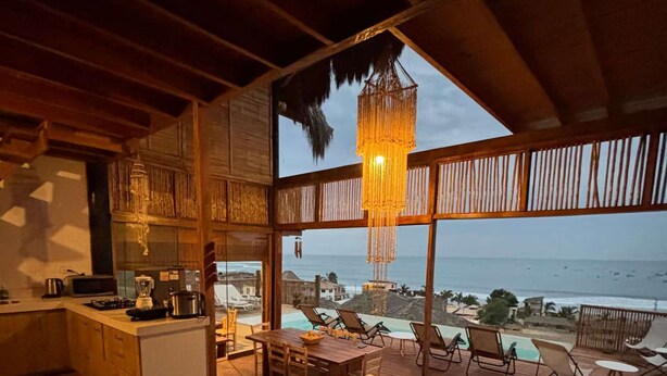 A spacious room with a large window overlooking the sea, a wooden dining area, and a kitchen with a bar. - w Relaxing Oceanfront 5BR in Punta Veleros (Los Organos)