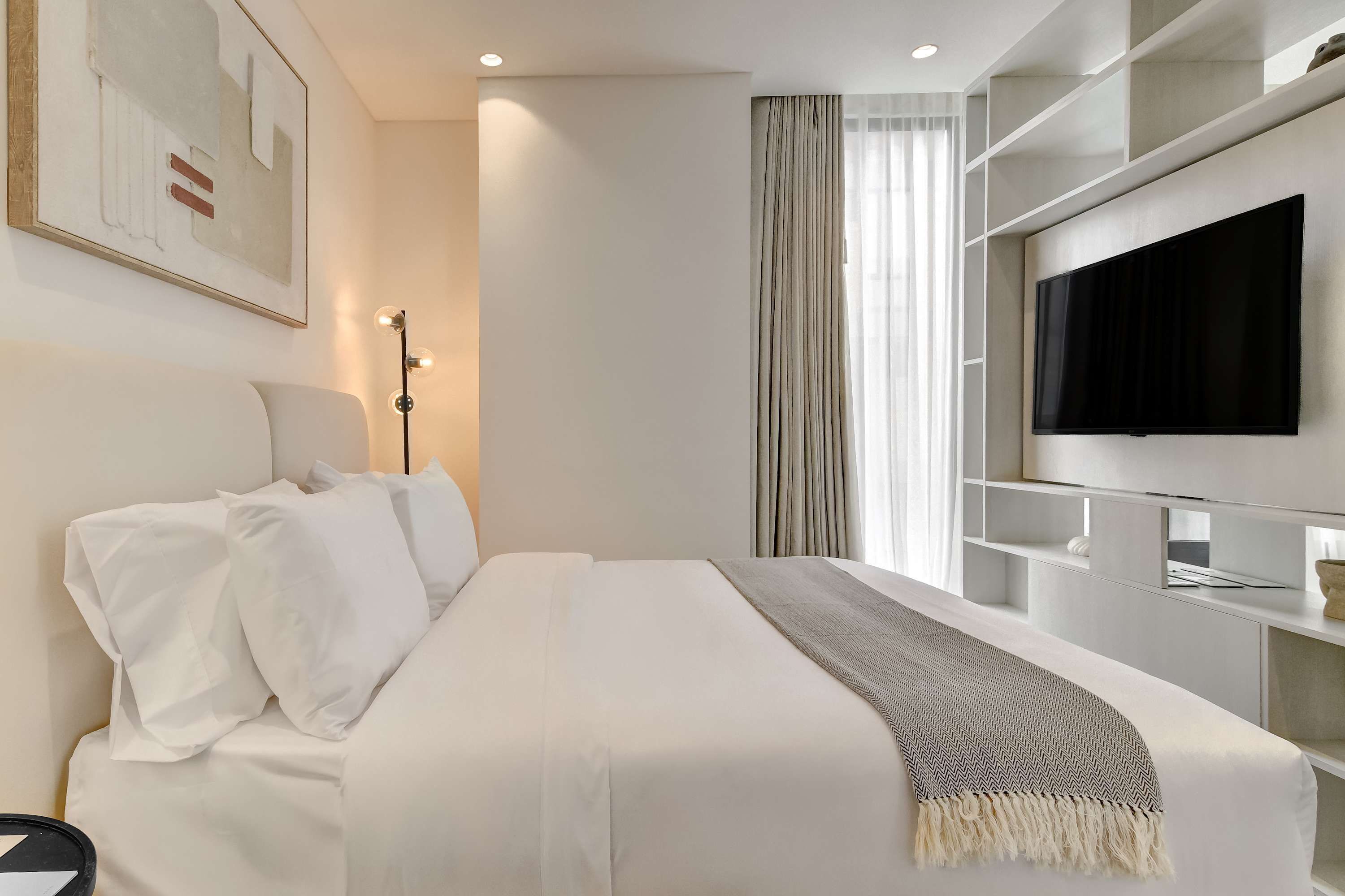 Habitación de hotel moderna con cama, televisor de pantalla plana en la pared, mesita de noche y un ventanal con cortinas.