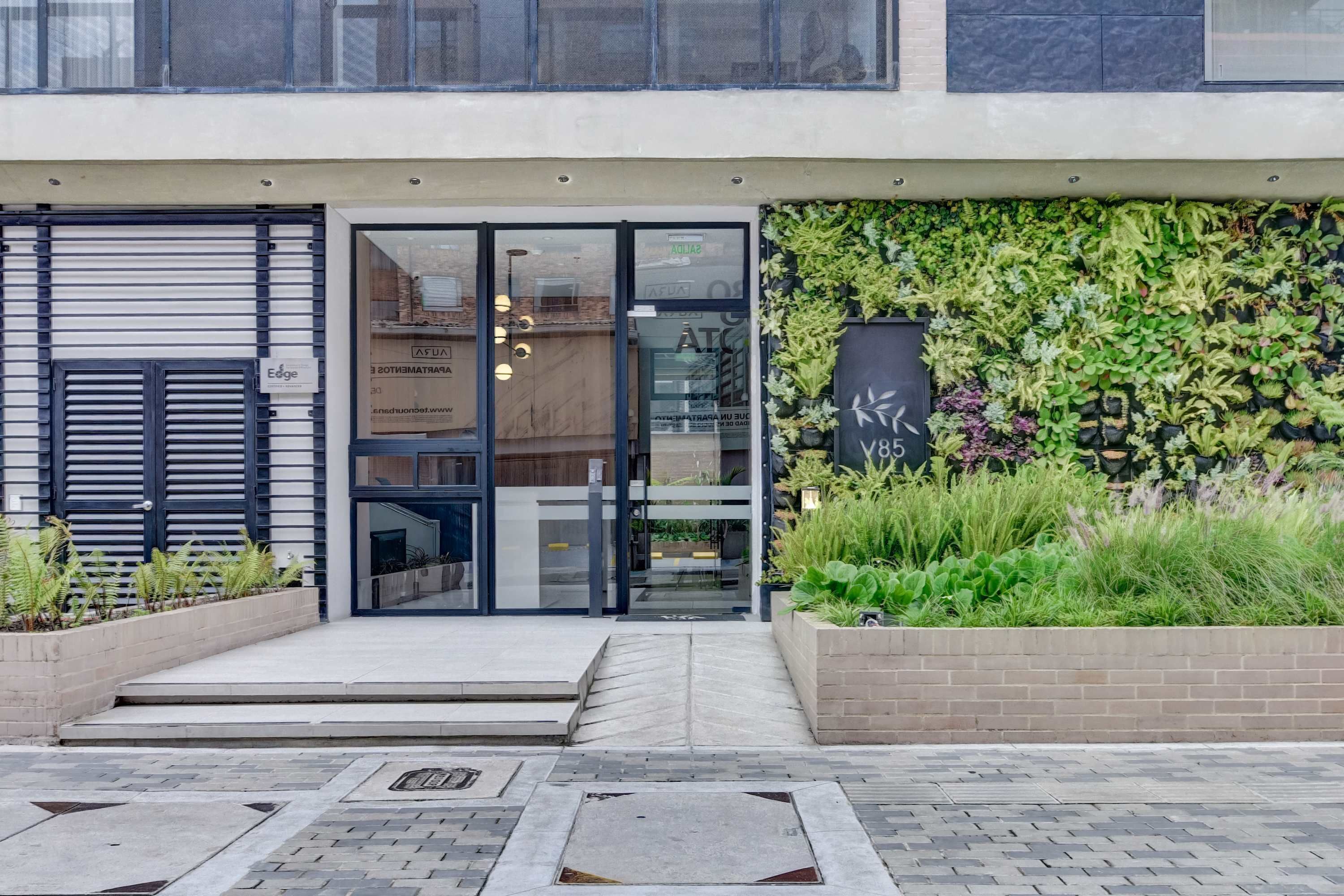 A modern building entrance with a glass door, a brick planter with greenery, and a signboard.