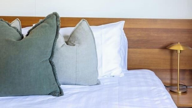 Quarto de hotel com cama, almofadas, abajur e duas pinturas emolduradas de paisagens na parede.