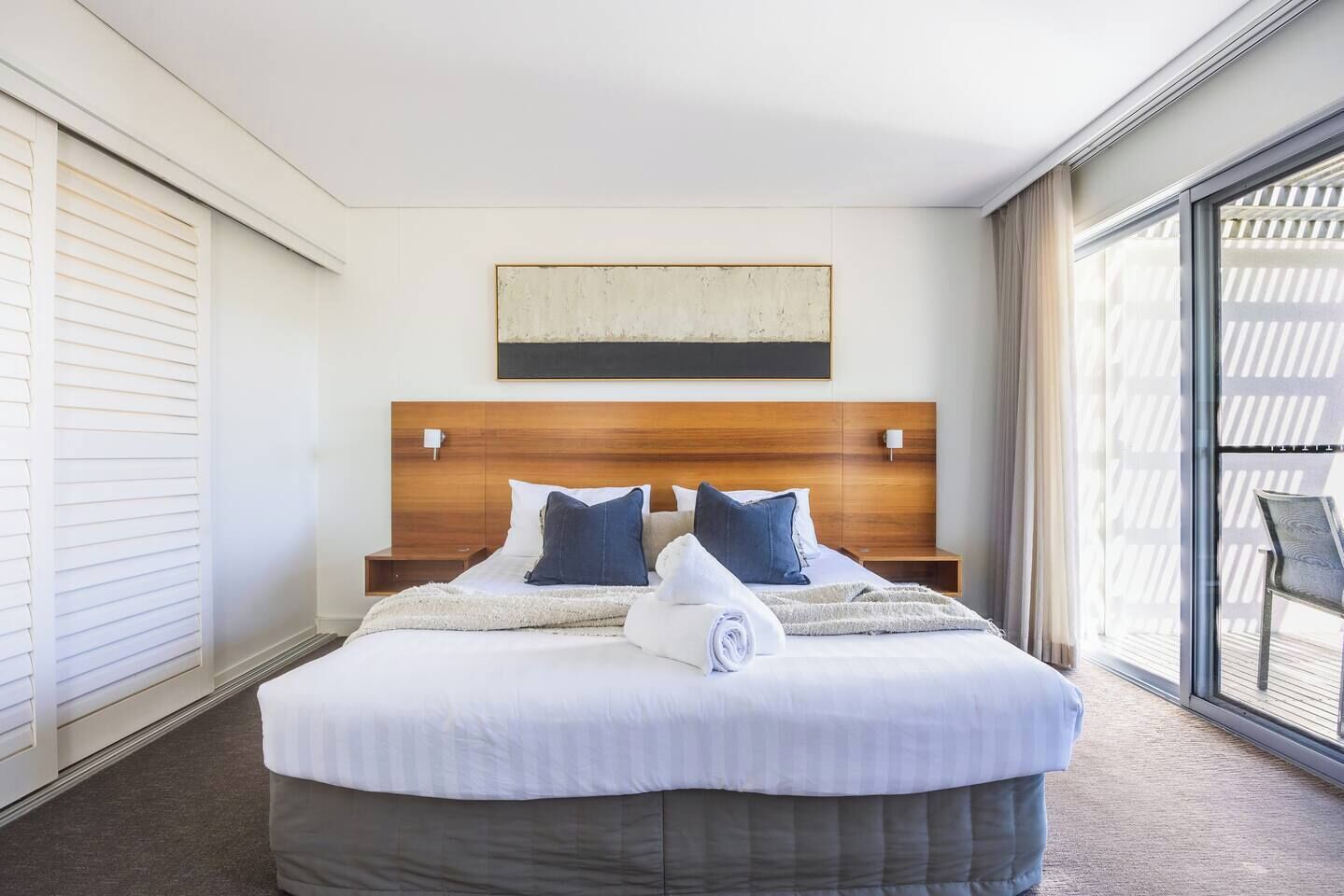 Ett hotellrum med en stor säng, ett träfärgat sänggavel, två kuddar, ett vitt överkast och utsikt genom ett stort fönster.