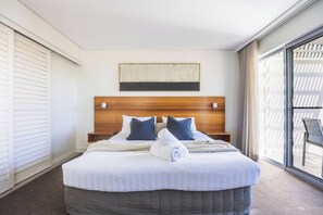 Ett hotellrum med en stor säng, ett träfärgat sänggavel, två kuddar, ett vitt överkast och utsikt genom ett stort fönster.