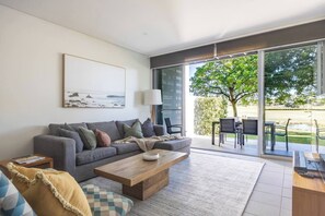 Ein modernes Wohnzimmer mit einem grauen Sofa, einem Holztisch und einem großen Fenster mit Blick auf einen Baum und Sitzmöglichkeiten im Freien.