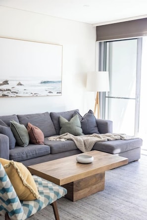 Ein modernes Wohnzimmer mit einem grauen Ecksofa, einem Holztisch und einem großen Fenster mit Blick ins Freie.