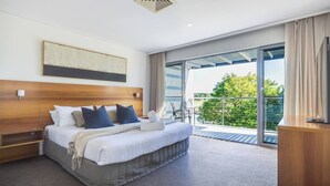 A modern hotel room with a large bed, a wooden headboard, a view of greenery through the balcony, and a ceiling vent.