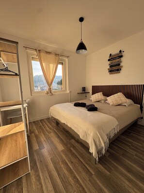Une chambre Ă coucher avec un lit, un plancher en bois, une fenĂȘtre avec des rideaux, un luminaire suspendu et un mur ornĂ© de plaques dĂ©coratives.