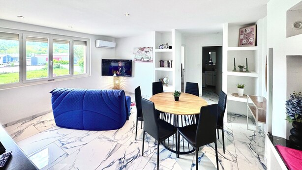 Uma sala de estar moderna com um puff azul, uma mesa de jantar de madeira com cadeiras, uma TV montada na parede e uma grande janela com vista para vegetação e edifícios.