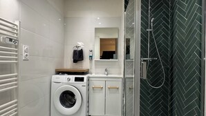 Um banheiro de hotel compacto com máquina de lavar, box de chuveiro em vidro e piso de mármore.