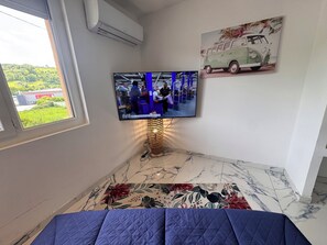 Une chambre dâhĂŽtel Ă©quipĂ©e dâune tĂ©lĂ©vision, dâune lampe dĂ©corative, dâun tapis Ă motifs floraux et dâun tableau accrochĂ© au mur.