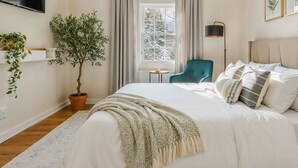 Ein Schlafzimmer mit einem Bett, einer Zimmerpflanze, einem Sessel, einem Fenster mit Vorhängen und einem an der Wand befestigten Fernseher.