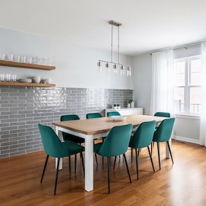 A modern kitchen with a brick backsplash, wooden shelves, teal chairs, and a hanging light fixture.