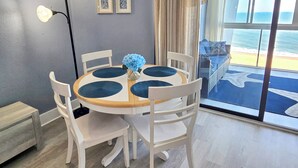 En matplats med ett runt bord, vita stolar, en vas med blå blommor och utsikt över stranden genom stora fönster.