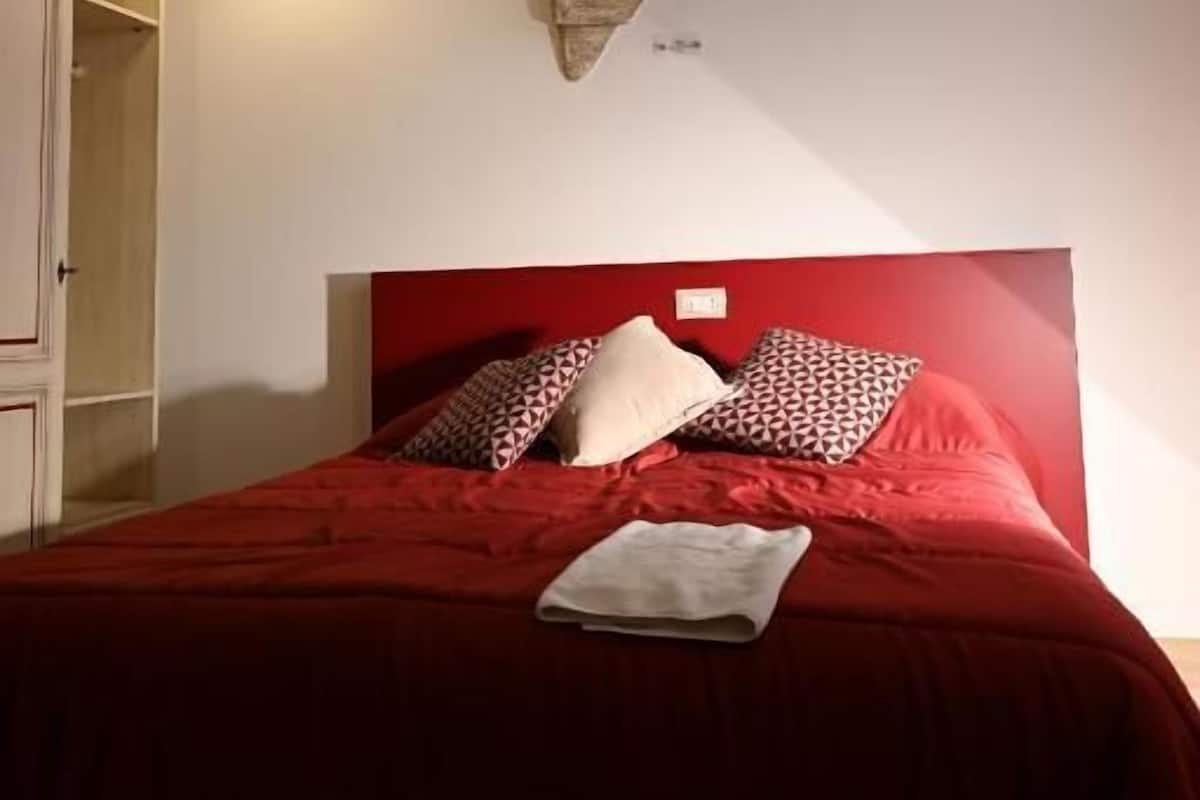 Una habitación con cama roja, pared de piedra y techo de madera.