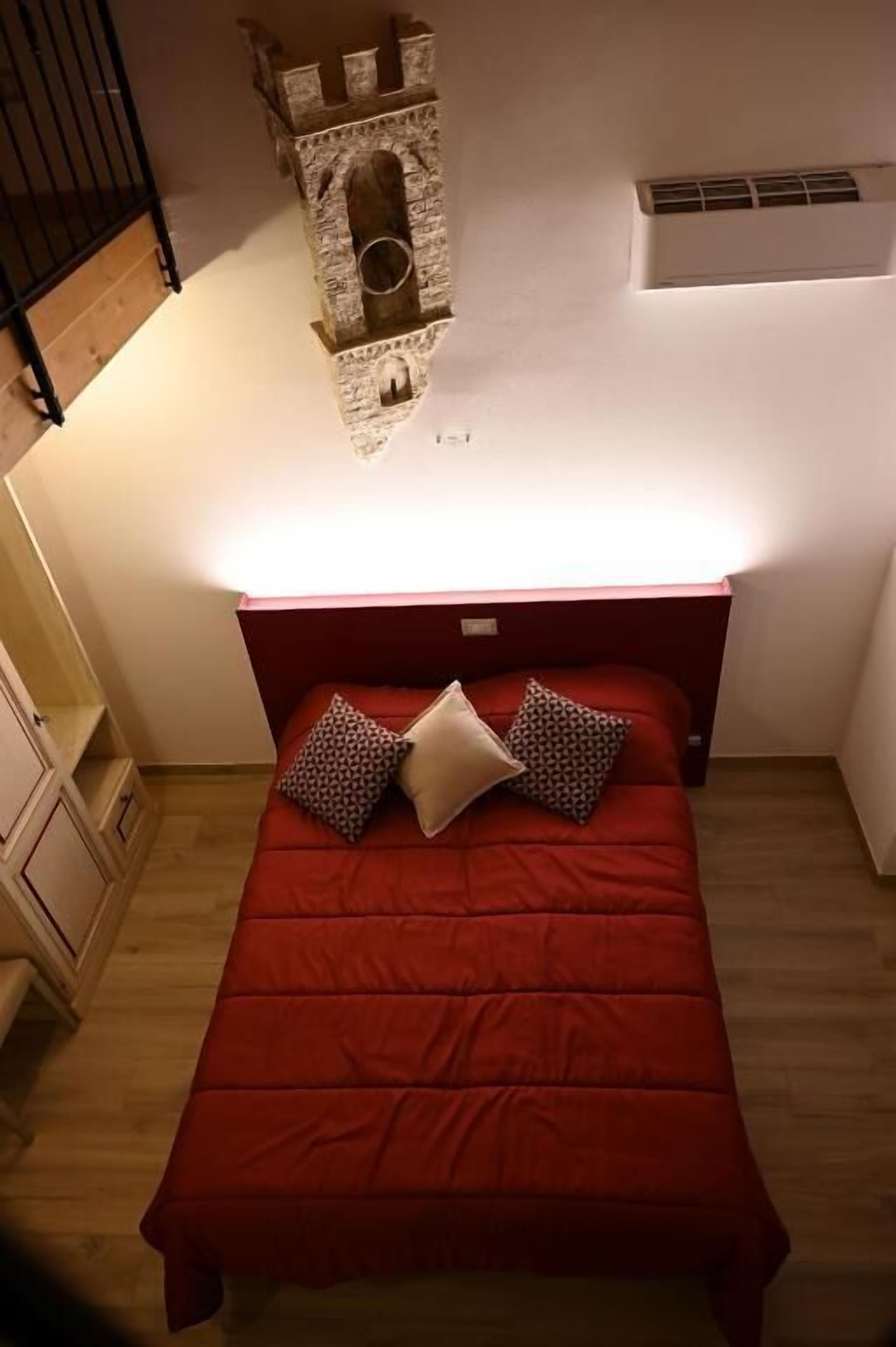 A bedroom with a red bedspread, two pillows, a stone tower-like structure on the wall, and a wooden floor.