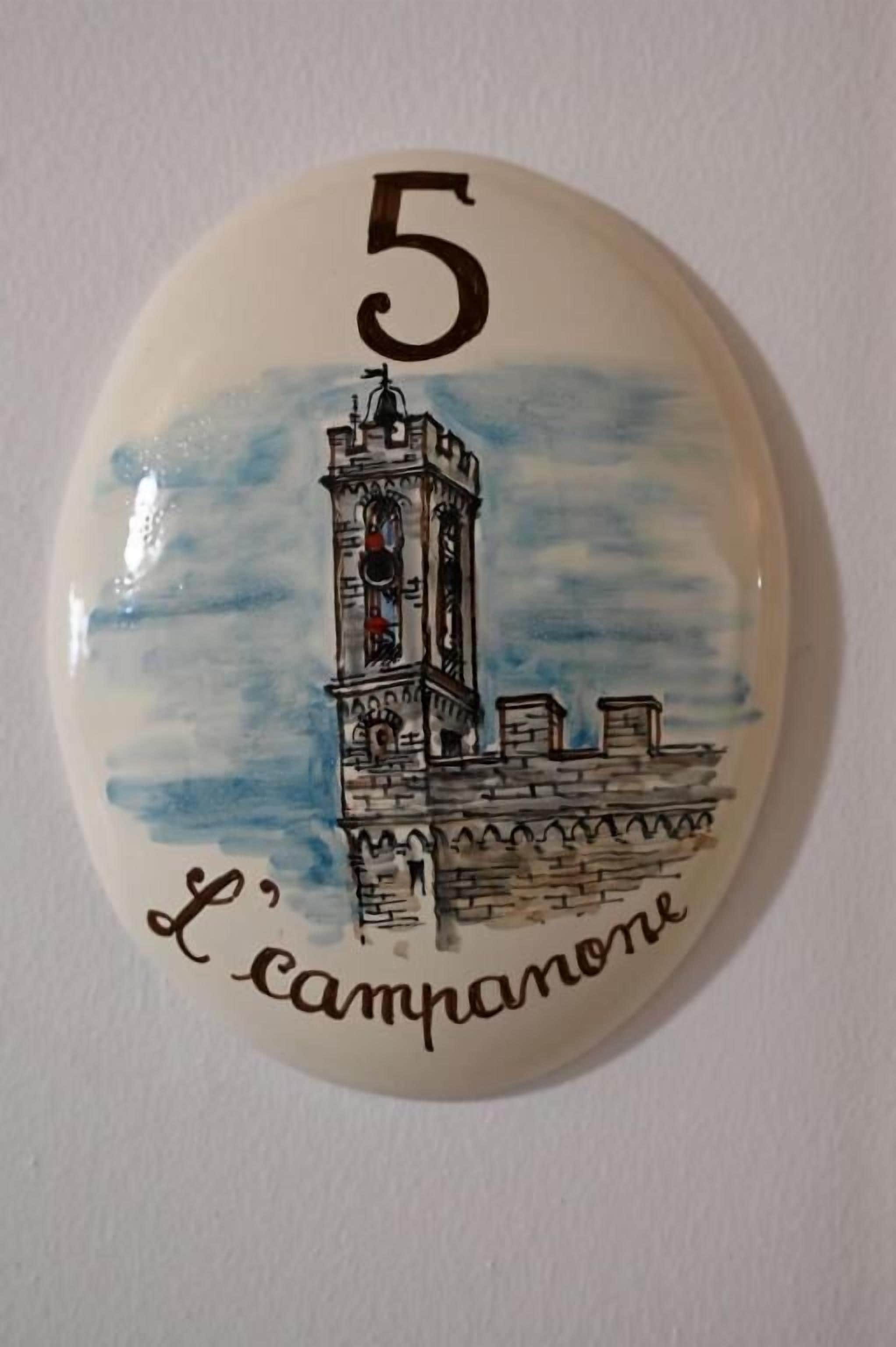 Una placa de cerámica con el número '5' y un dibujo de una torre, posiblemente un punto de referencia, con el texto 'L'campanone' debajo.