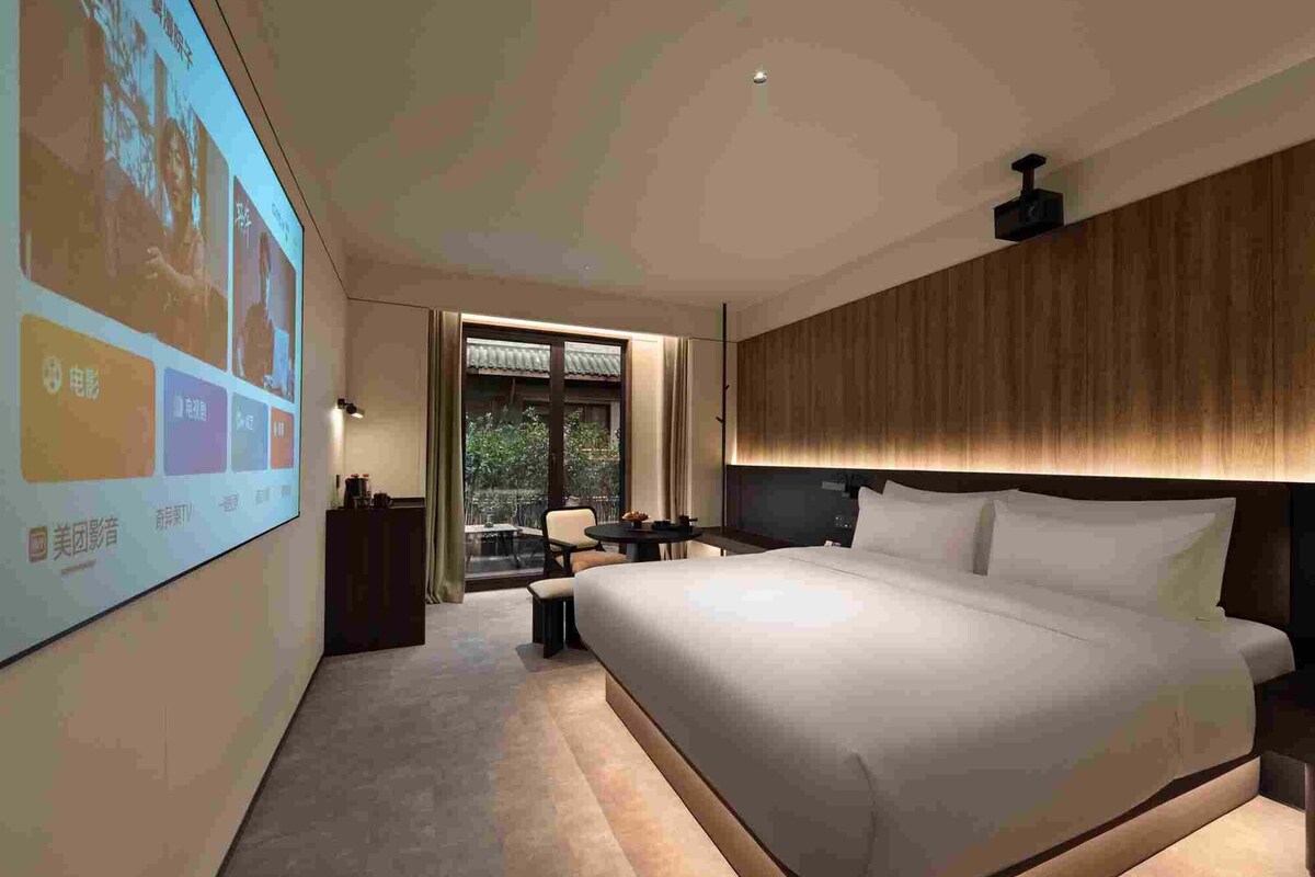 A modern hotel room with a large bed, a desk, a chair, and a projection screen on the wall.