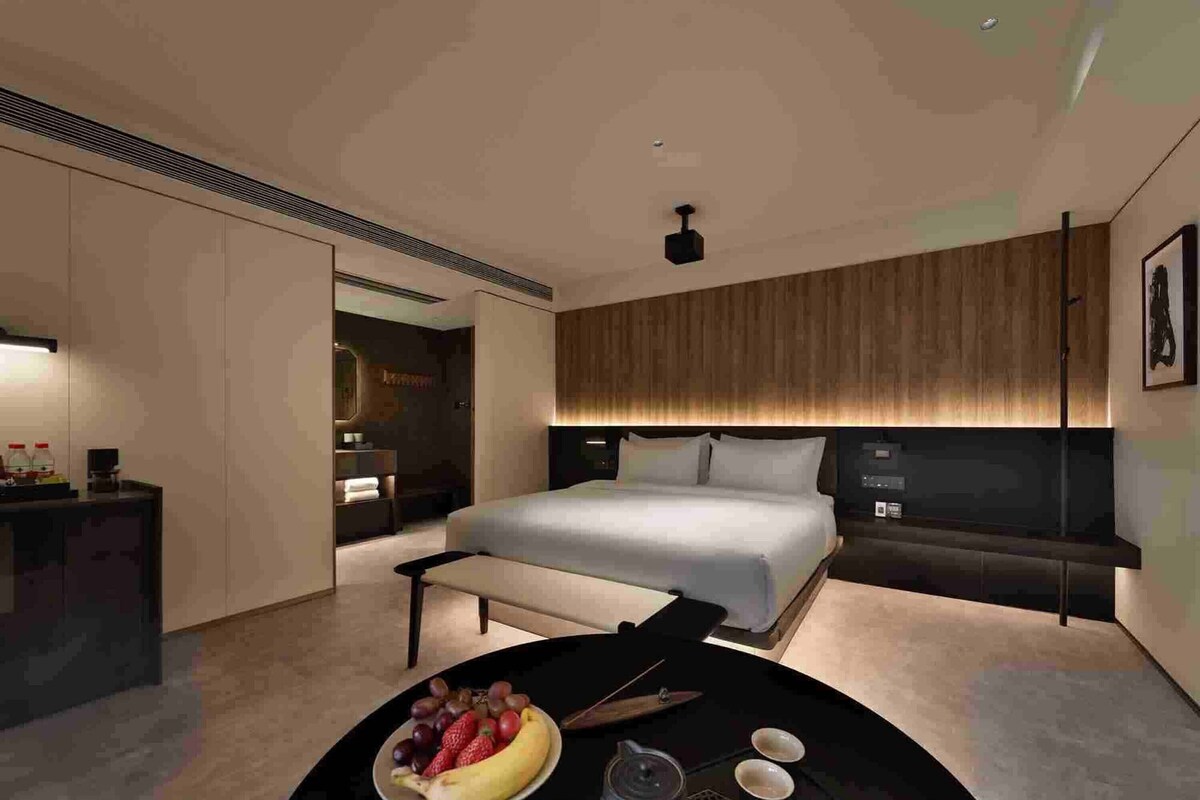 A modern hotel room with a large bed, a bedside table with a fruit bowl, a coffee maker, and two cups.