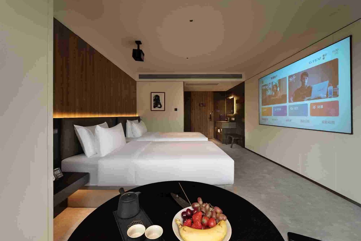 A modern hotel room with a bed, a desk with a laptop, a fruit bowl, and a projection screen displaying a presentation.