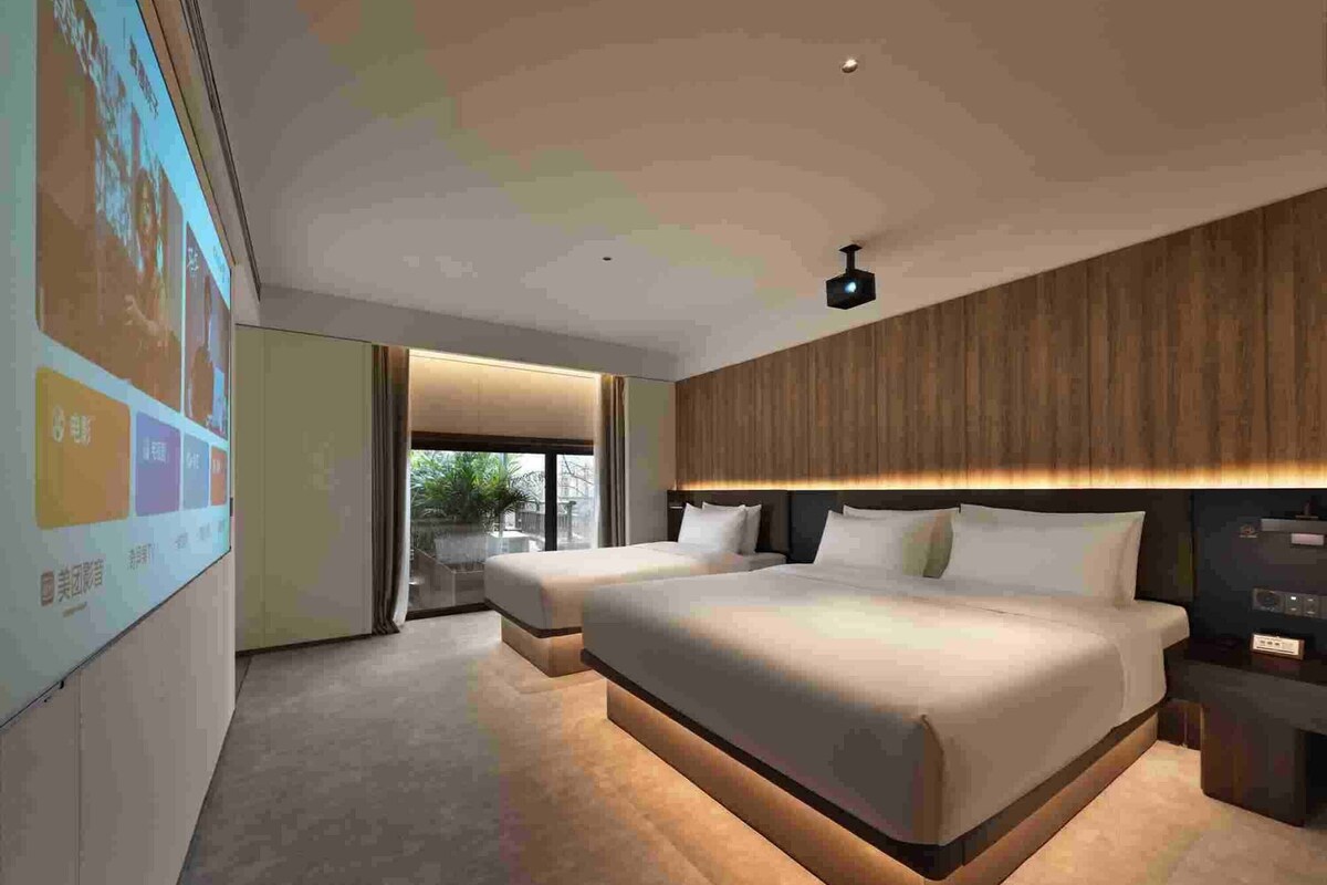 A modern hotel room with two beds, a large window, and a projection screen on the wall.