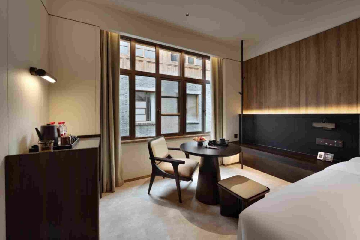 A hotel room with a bed, a desk with a lamp, a chair, a round table, and a large window with a view of buildings.